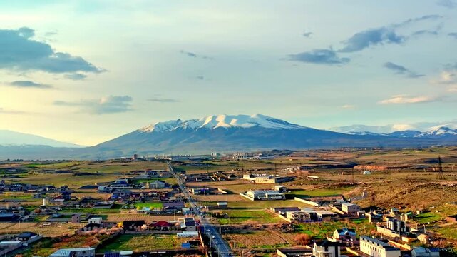 Panoramic view of town nestled in valley before snow-capped mountain. Sky has soft clouds, light filters through. Landscape features green fields and scattered homes