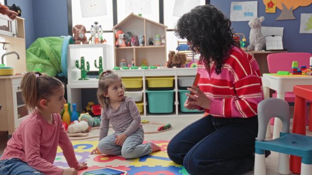 Teacher kneels showing a flashcard to two girls on a colorful foam mat in a preschool classroom building; playful learning.