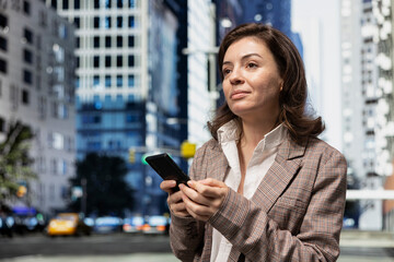 Fototapeta premium Close up of smiling woman holding smartphone traversing city blocks to reach office building. Cheerful businesswoman walking on metropolitan avenue to job location, using mobile phone