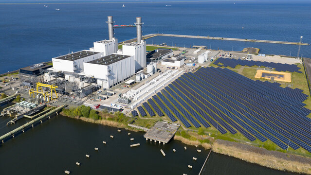 Aerial view of the ENGIE Maxima power plant featuring white industrial buildings, tall chimneys, and a large solar panel array along the coast in Lelystad, Flevoland, Netherlands.