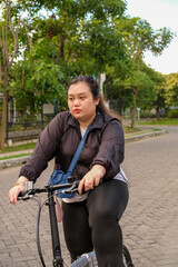 Obraz premium Woman riding a bicycle outdoors, looking thoughtful, with trees and a paved path in the background.