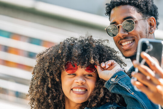 young man and woman laugh and pose for a selfie outside. They wear sunglasses and share a fun, joyful moment together