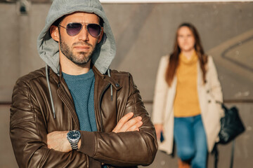 confident man in a leather jacket, hood, and sunglasses stands with arms crossed on a urban street, with a woman walking in the background © carballo