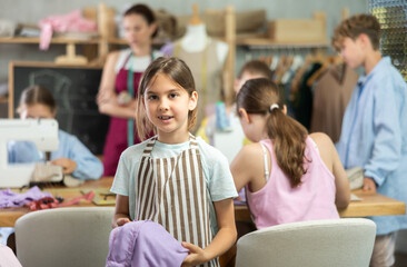 Obraz premium Positive girl child in study room of sewing school and young designers. Group of students in background during discussion of work processes