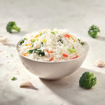 A bowl of steamed white rice mixed with colorful vegetables including broccoli, bell peppers, and leafy greens, served on a light textured surface with scattered gar