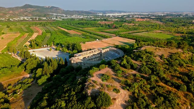 Wide drone view of Aspendos Theatre landscape. Ancient amphitheater sits in the Turkish countryside among fields, trees and distant hills.