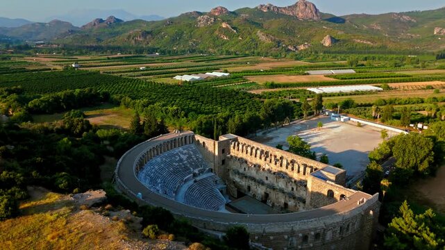 Drone orbit over Aspendos Theatre in Turkey. Ancient Roman amphitheater is seen from above with rural fields and mountains nearby.