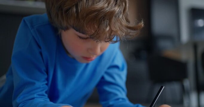 Young boy in blue shirt concentrating as he writes or draws with a pencil on paper at a desk in a softly lit indoor learning environment