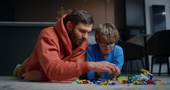 Father and young son lying on the floor assembling colorful toy building bricks together, sharing focused playtime in a cozy indoor room
