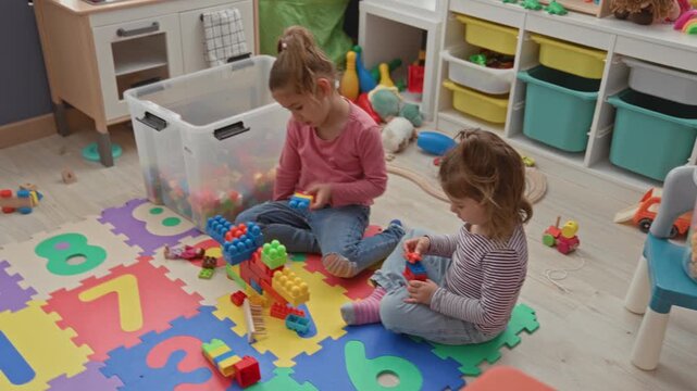 Two girls build a colorful block tower on a foam number mat in a playroom, one stacking bricks while the other holds pieces; quiet concentration.