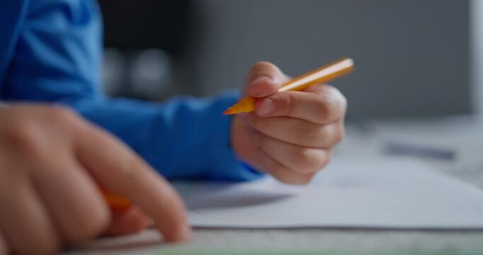 Close-up of a child in a blue sleeve drawing with a yellow pencil on white paper at a table, showing focused hands, learning, creativity, and early education