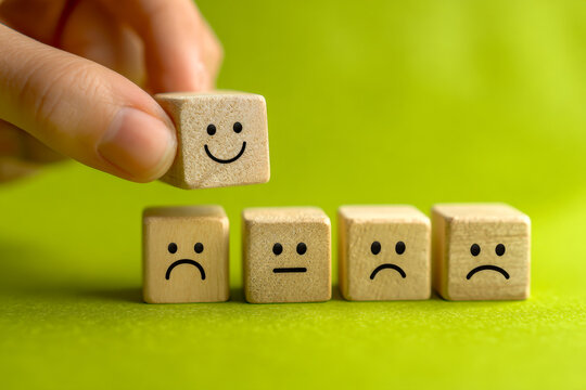 Hand placing a wooden block with a happy face among sad and neutral face blocks symbolizing positivity and emotional change concept on green background
