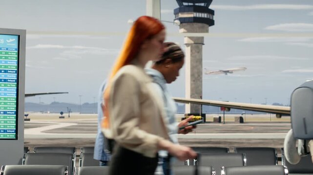 Tourists commuting in airport transport hub waiting area overlooked by runway control tower. People walking through rows of chair seats inside bright modern flight departure terminal, camera A