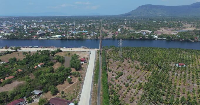 Backward aerial view showing a road intersection, buildings, and palm plantation in the foreground, leading to a freight train crossing a river bridge with a town and mountains in the background.