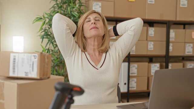 Woman with hands behind head and closed eyes amid stacked packages, laptop and scanner in a packing area inside a building; relief.