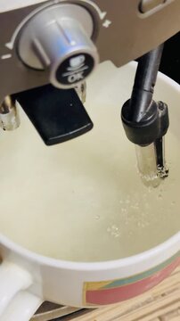 Steam wand rinsing white coffee mug during descaling cycle, hot water and foam flow from stainless nozzle into mug, foamy bubbles