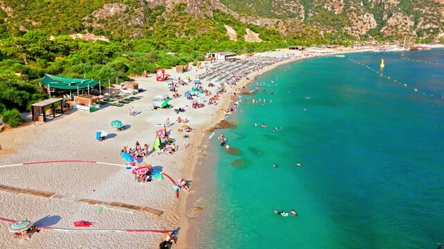 Drone view of Oludeniz Blue Lagoon, Turkey with umbrellas. People relax on the sandy beach beside turquoise water and green mountains.