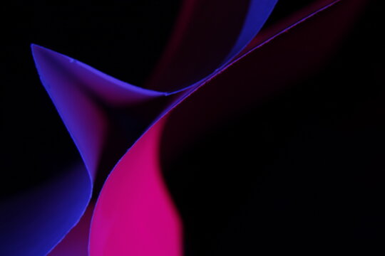 Composici&oacute;n abstracta de papeles coloridos con formas curvas y triangulares iluminadas con luces ne&oacute;n sobre fondo negro. La combinaci&oacute;n de tonos magenta, azul y violeta crea un efecto visual moderno.