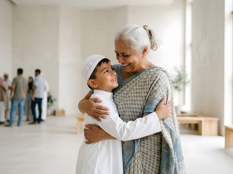 Happy grandmother in traditional sari hugging her grandson wearing white thobe and peci in bright hall