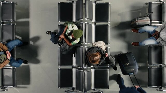Diverse passengers walking in busy airport departure terminal, sitting down on chairs, top down view. Multiracial people waiting on benches to fly home during holiday rush, high angle shot
