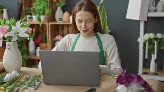 Woman florist with green apron closes a laptop with both hands at a flower shop counter surrounded by vases and lilies in a studio; small business warmth.