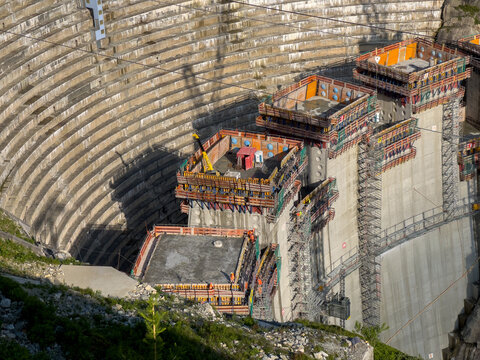 Aerial view of a massive concrete dam under construction with scaffolding, industrial platforms, and workers active. The Dam is an artificial lake near the Grimsel Pass, Canton of Bern