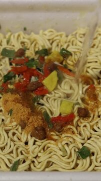 Vertical video close up. Hot boiling water from kettle is poured in plastic plate bowl with ramen, quick cooking instant egg curly noodles with herbs, spices for Asian soup. Healthy fast food, lunch