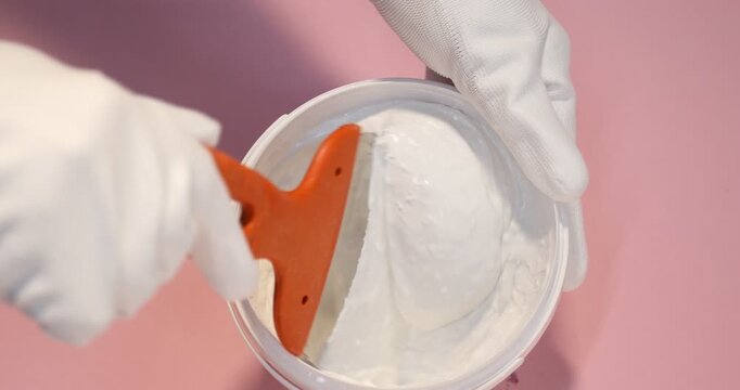 Worker with putty knife and bucket of plaster on pink background, top view