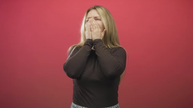 Woman with hand covering mouth, black shirt and jeans, centered against red studio backdrop; distress vulnerability.