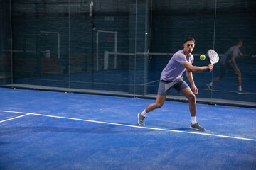 Obraz premium Two male players lunging swinging padel racket at greenball in purplepolo on blue court, copy space