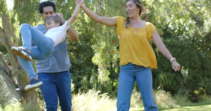 Diverse family parents bending, lifting and swinging child by hands on grassy lawn, smiling
