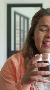 Vertical video: Bringing mug to lips, woman in peach top sipping, wincing from hot drink in kitchen