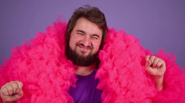 Horizontal 4K video. Mature man with beard, glitter sparkles heart to his cheek, face, pink magenta tulle fur coat, purple t-shirt smiles, dances, looks at camera. Birthday holiday. Pride lgbt month