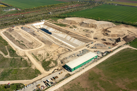 Brasov, Romania - 28 April 2026: Aerial view of a waste management facility with a large excavation pit, industrial buildings, and heavy machinery surrounded by green fields.