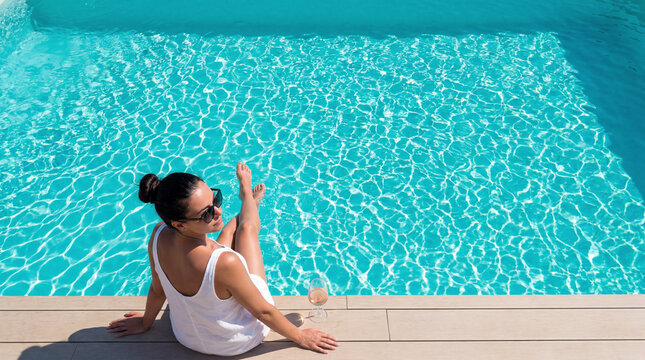 Description: High-angle view of a woman in white summer attire relaxing by a turquoise swimming pool with a glass of wine on a sunny day.