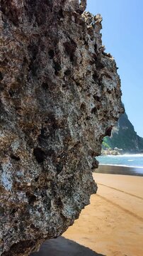 Cinematic reveal of a beautiful sandy beach with blue ocean waves and green cliffs from behind a coral rock. Tropical paradise landscape on a sunny day. Parangtritis, Yogyakarta, Indonesia