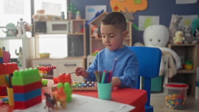 Hispanic boy seated at table plays xylophone with mallet using his hand among toys in a preschool building; concentration learning.