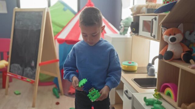 Hispanic boy holding green plastic robot toy while leaning and reaching with hand over shelf in building playroom scattered with toys and a chalkboard; curiosity.