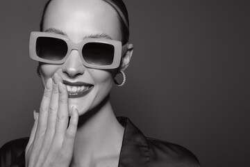 Fototapeta premium Smiling young white woman with pale skin wearing large square sunglasses and hoop earrings covers mouth with hand. Black and white studio portrait with elegant mood and fashion style.