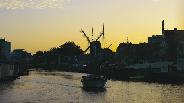 Silhouette of traditional windmill beside calm canal during golden sunset as small boat moves across water creating peaceful reflection in scenic Dutch landscape
