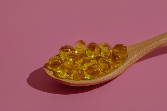 Yellow soft gel capsules on a wooden spoon casting shadow against a pink background symbolizing vitamins and dietary supplements for health and wellness