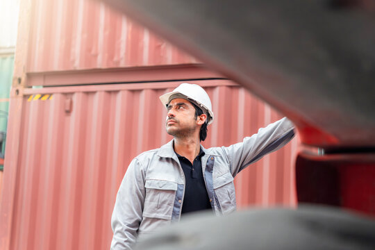 Logistics worker in hard hat and uniform inspecting cargo at container terminal, Engineer standing by red shipping containers in a maritime port, Foreman worker overseeing operations in a shipyard