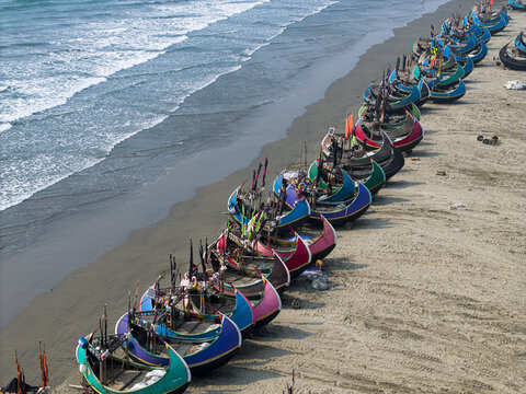 Cox's Bazar, Bangladesh - 10 March 2026: Aerial view of a long row of colorful traditional moon boats parked on the sandy shore of the beach with people and ocean waves crashing nearby.