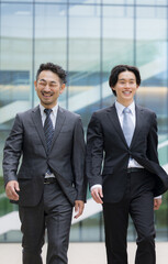 Fototapeta premium 都会のビル背景で笑顔で横並びで歩くハンサムな営業チームのポートレート、法人営業や企業成長など若手から管理職のポジティブイメージ