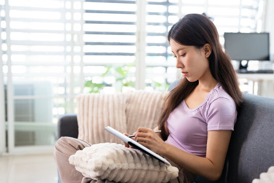 Young Asian woman studying online, sitting on sofa with teblet at home.