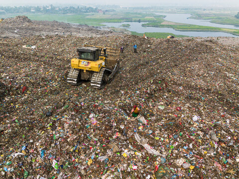Aminbazar, Bangladesh - 26 January 2025: Aerial view of a yellow bulldozer moving waste at the Aminbazar landfill with workers scavenging among piles of garbage near a river.