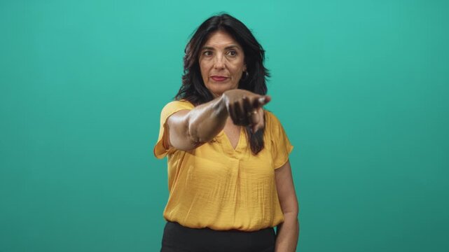 Woman pointing finger forward in studio wearing yellow blouse against a teal backdrop; confidence command.