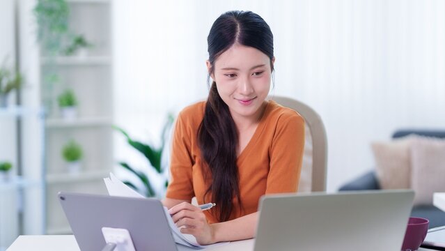 Asian businesswoman working from home office using laptop computer writing document hybrid work remote professional sitting desk modern apartment workspace