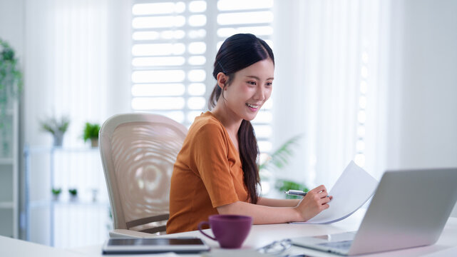 Asian woman working from home office writing document reading paperwork using laptop computer sitting desk bright modern workspace remote hybrid work freelance