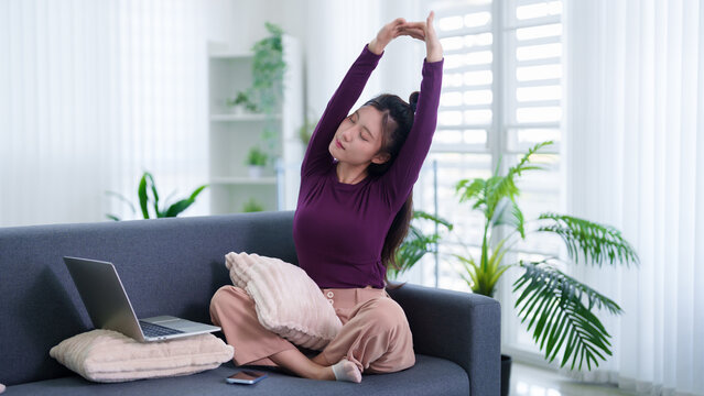 Asian women doing stretching exercise work from home sitting on sofa with laptop computer relaxing wellness routine during remote working break at modern